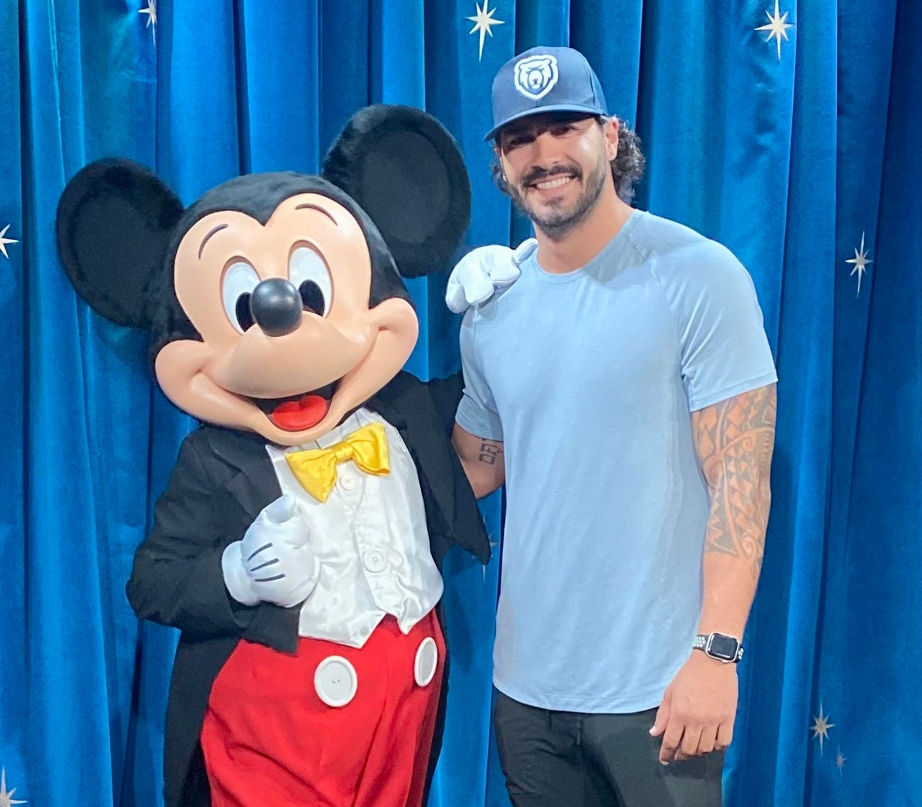 mickey mouse places a gloved hand on a smiling man in a blue t-shirt and baseball cap